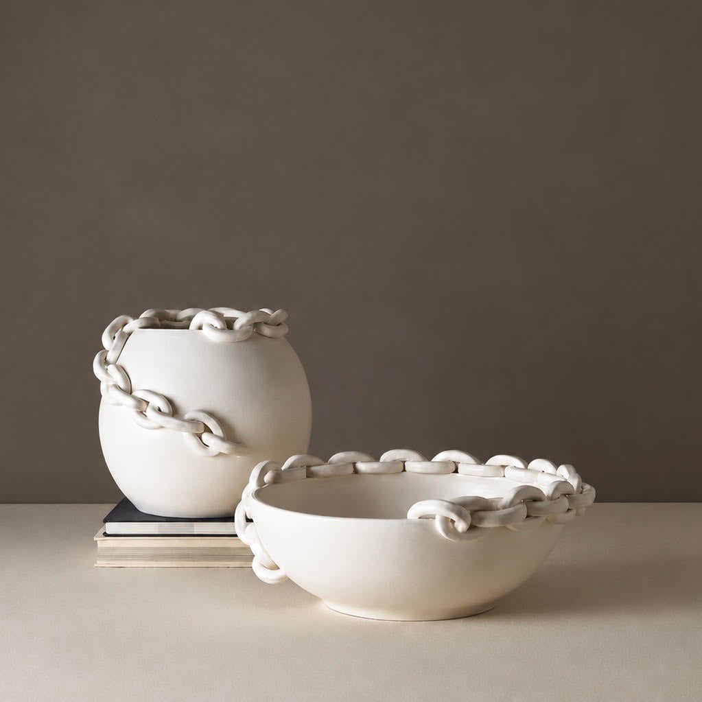 Two matte white ceramic decorative pieces, a round vase and a shallow bowl, both featuring sculptural chain-like handles around the rims, displayed on a neutral beige surface against a taupe background.