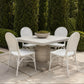 A white wicker and metal patio dining set with a square stone-look table and four matching chairs, arranged on a stone patio in front of tall green hedges.