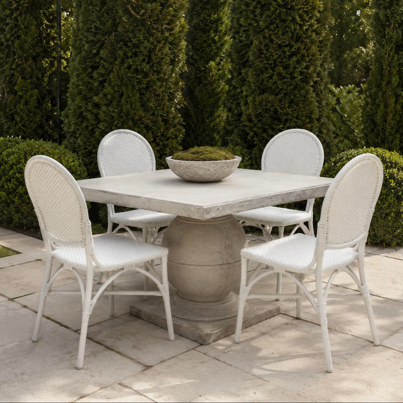A white wicker and metal patio dining set with a square stone-look table and four matching chairs, arranged on a stone patio in front of tall green hedges.
