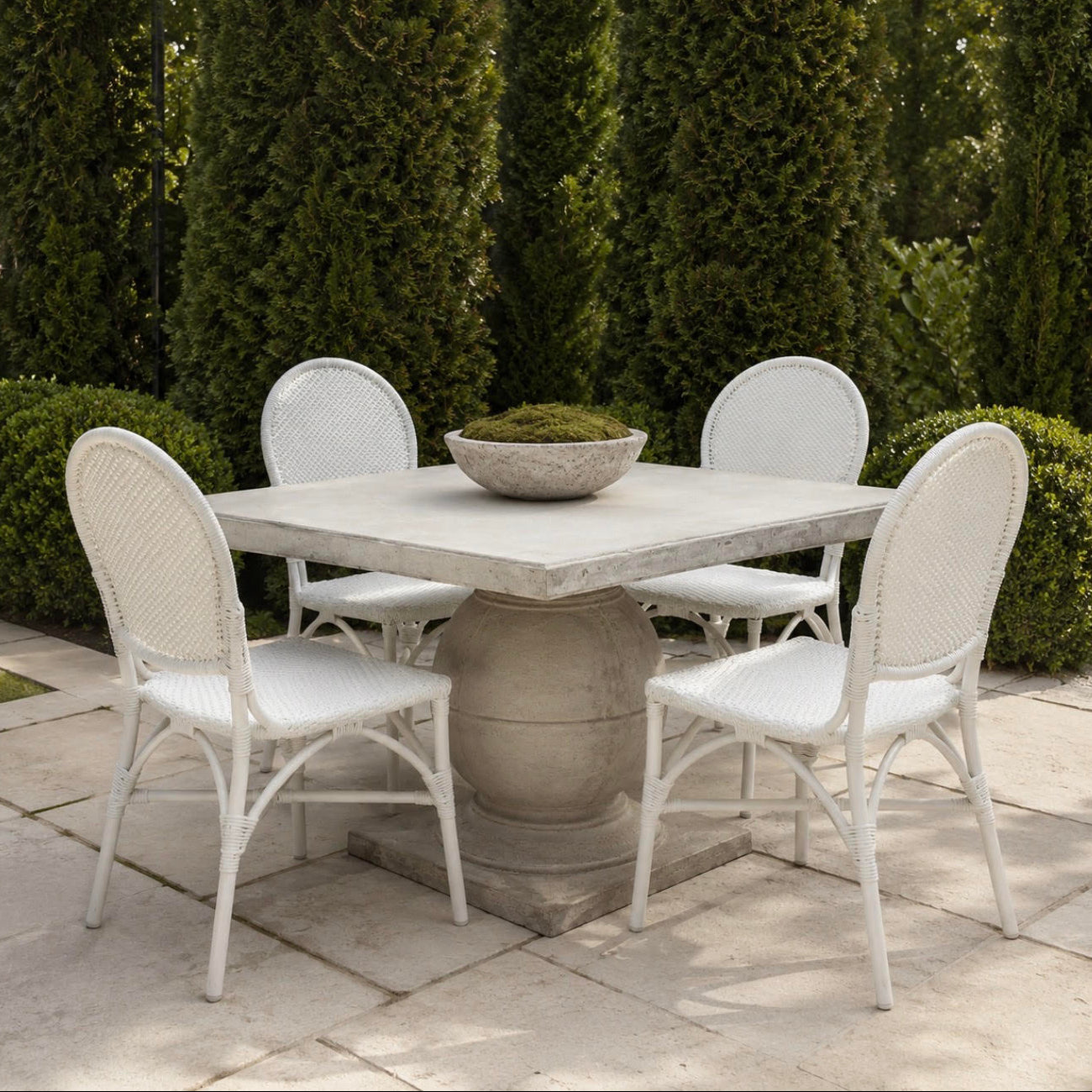 A white wicker and metal patio dining set with a square stone-look table and four matching chairs, arranged on a stone patio in front of tall green hedges.
