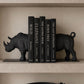 A set of six black hardcover books displayed on a beige shelf, flanked by two black rhinoceros bookends. The books are titled: WILD, ELEPHANT, THE LIVING WILD, BORN FREE, THE LAST ELEPHANTS, and THE KINFOLK HOME.