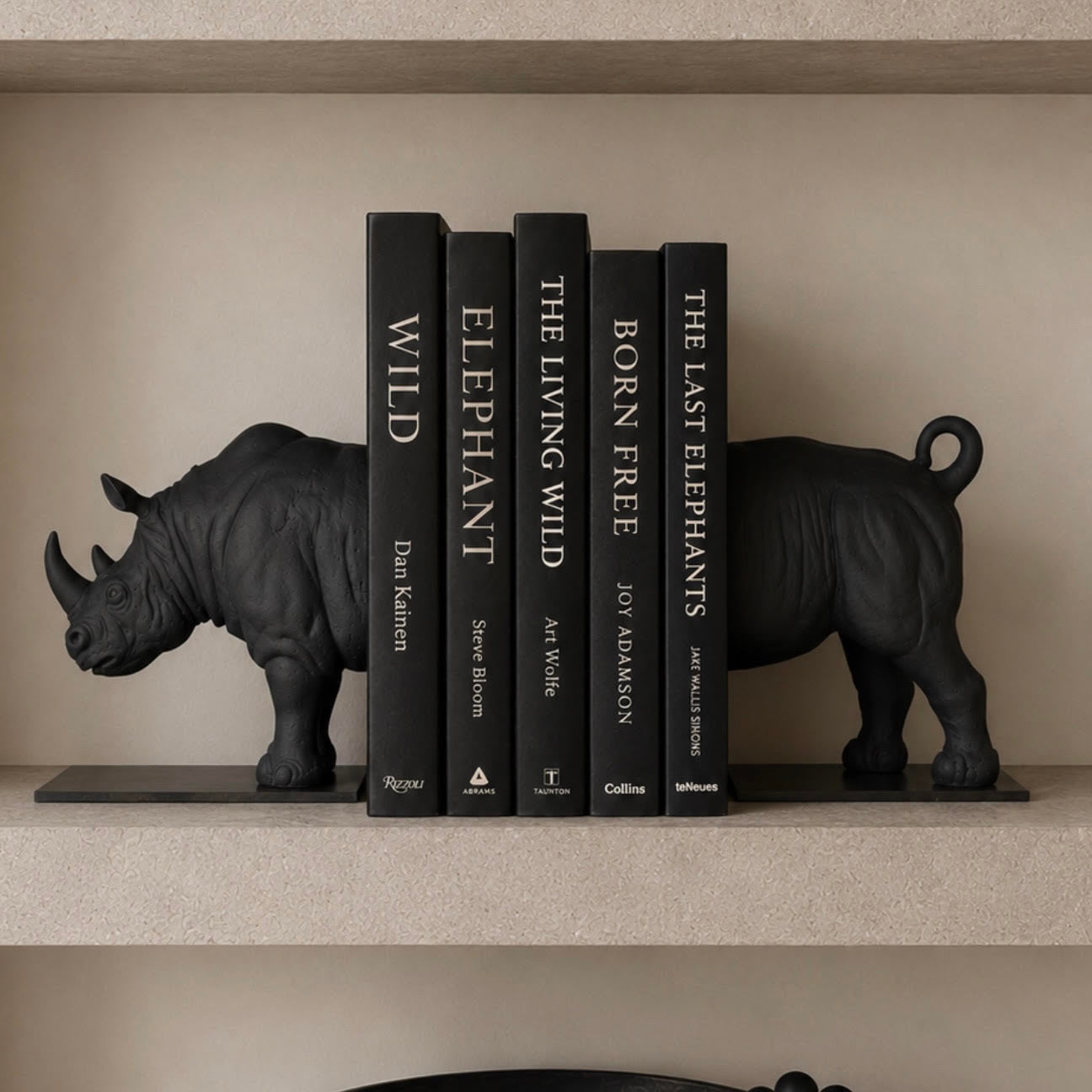 A set of six black hardcover books displayed on a beige shelf, flanked by two black rhinoceros bookends. The books are titled: WILD, ELEPHANT, THE LIVING WILD, BORN FREE, THE LAST ELEPHANTS, and THE KINFOLK HOME.