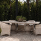 Outdoor dining set featuring a rectangular stone table with four wicker armchairs, arranged on a gravel patio with a backdrop of green hedges and trees.