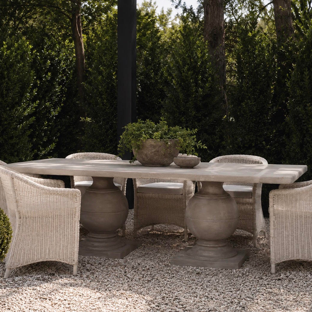 Outdoor dining set featuring a rectangular stone table with four wicker armchairs, arranged on a gravel patio with a backdrop of green hedges and trees.