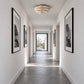 A long, bright hallway with white walls, framed black-and-white wall art, a textured ceiling light, and a dark stone floor leading to an open doorway.