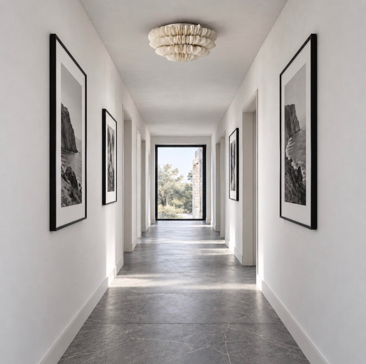 A long, bright hallway with white walls, framed black-and-white wall art, a textured ceiling light, and a dark stone floor leading to an open doorway.