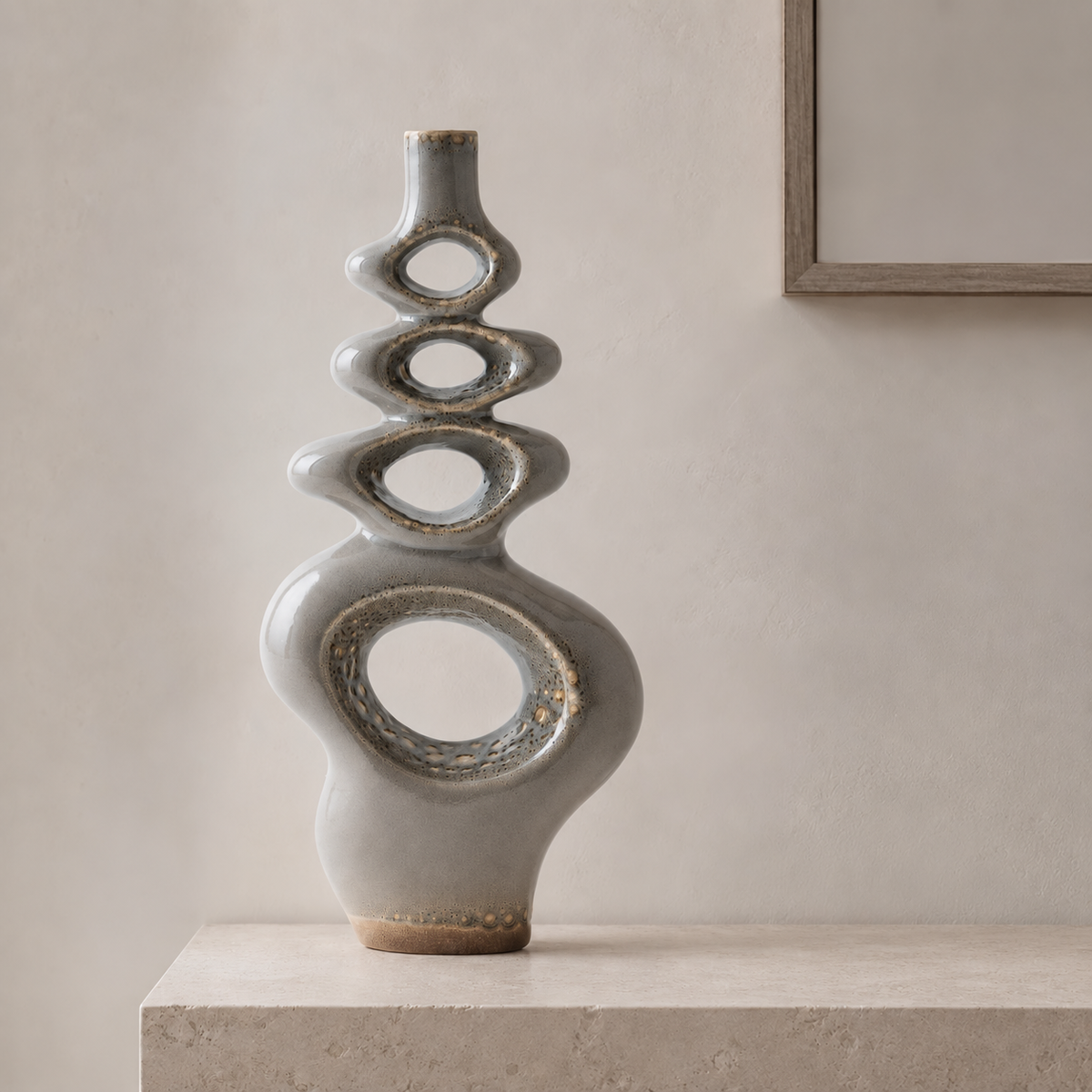 A tall, sculptural ceramic vase with a glossy gray-green glaze and organic open-ring shapes, displayed on a stone pedestal against a neutral beige wall.