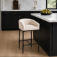 A modern kitchen island with a black cabinet base, white marble countertop, and a cream upholstered bar stool in the foreground. The kitchen features black cabinets, a white marble backsplash, a gold faucet, and a black pendant light.