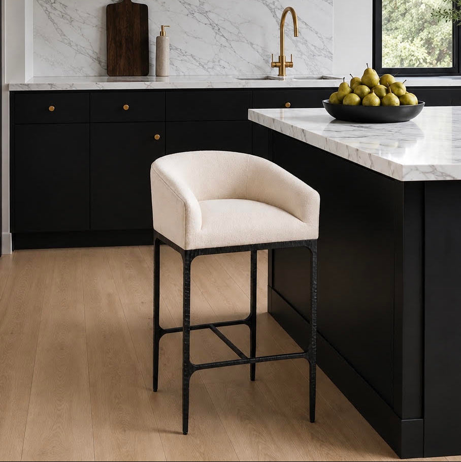 A modern kitchen island with a black cabinet base, white marble countertop, and a cream upholstered bar stool in the foreground. The kitchen features black cabinets, a white marble backsplash, a gold faucet, and a black pendant light.