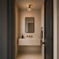 A modern bathroom vanity featuring a floating beige stone sink, a black-framed rectangular mirror, and brass fixtures, viewed from an open dark doorway with a warm ceiling light above.