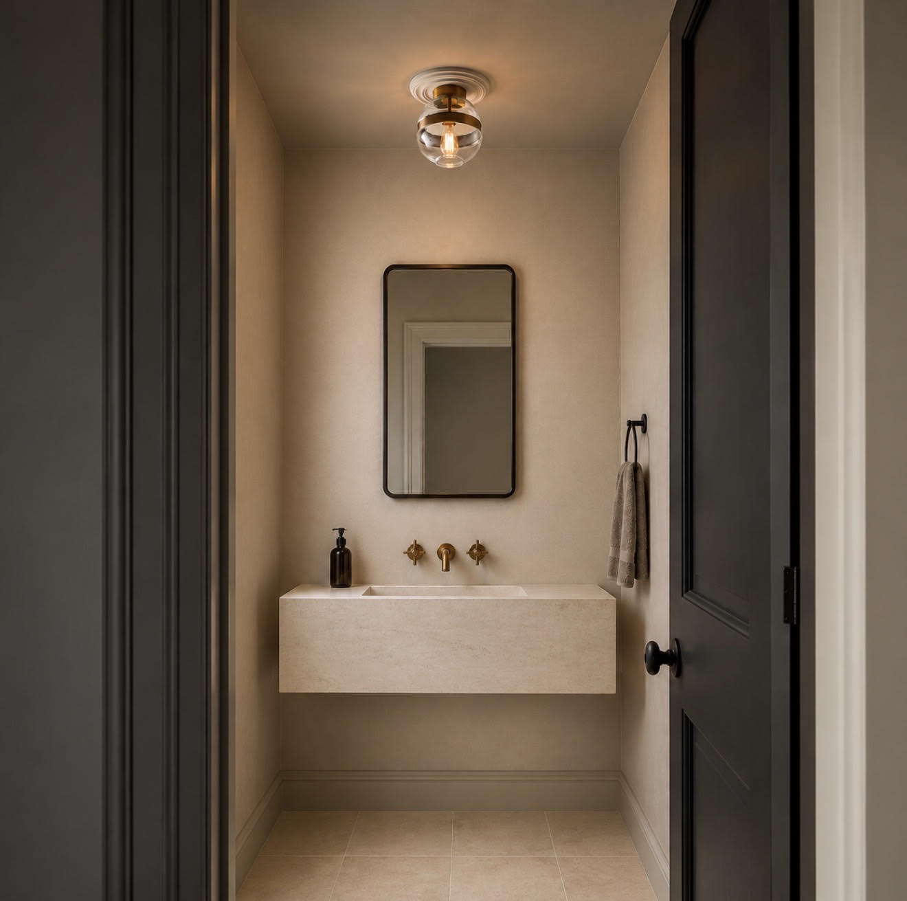 A modern bathroom vanity featuring a floating beige stone sink, a black-framed rectangular mirror, and brass fixtures, viewed from an open dark doorway with a warm ceiling light above.