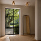 A modern interior room with two brass ceiling light fixtures, a tall floor mirror, and a large black-framed window looking out onto a garden with a tree.
