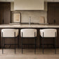 Three modern bar stools with cream upholstered seats and black metal frames, set in front of a kitchen island with a stone countertop and backsplash.