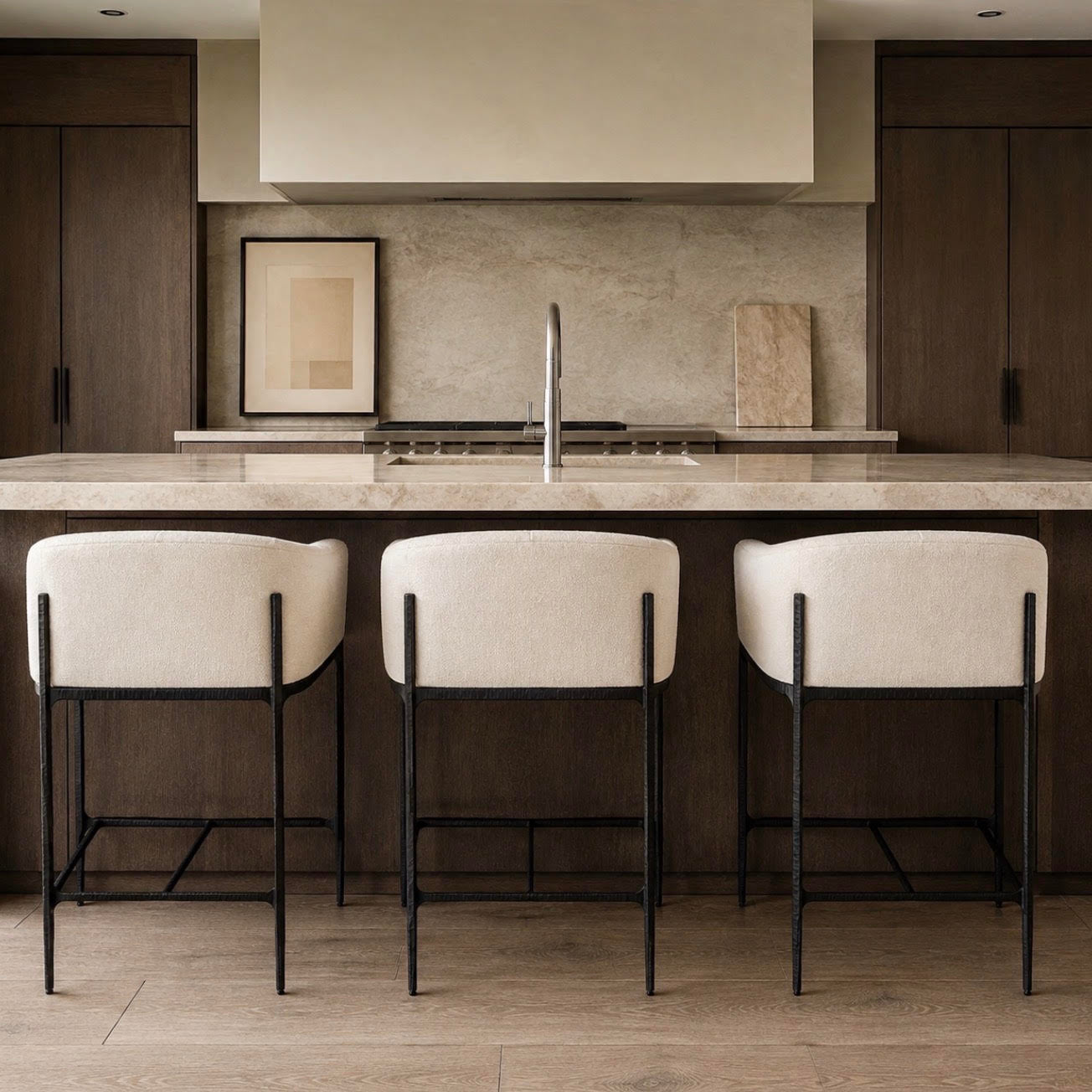 Three modern bar stools with cream upholstered seats and black metal frames, set in front of a kitchen island with a stone countertop and backsplash.