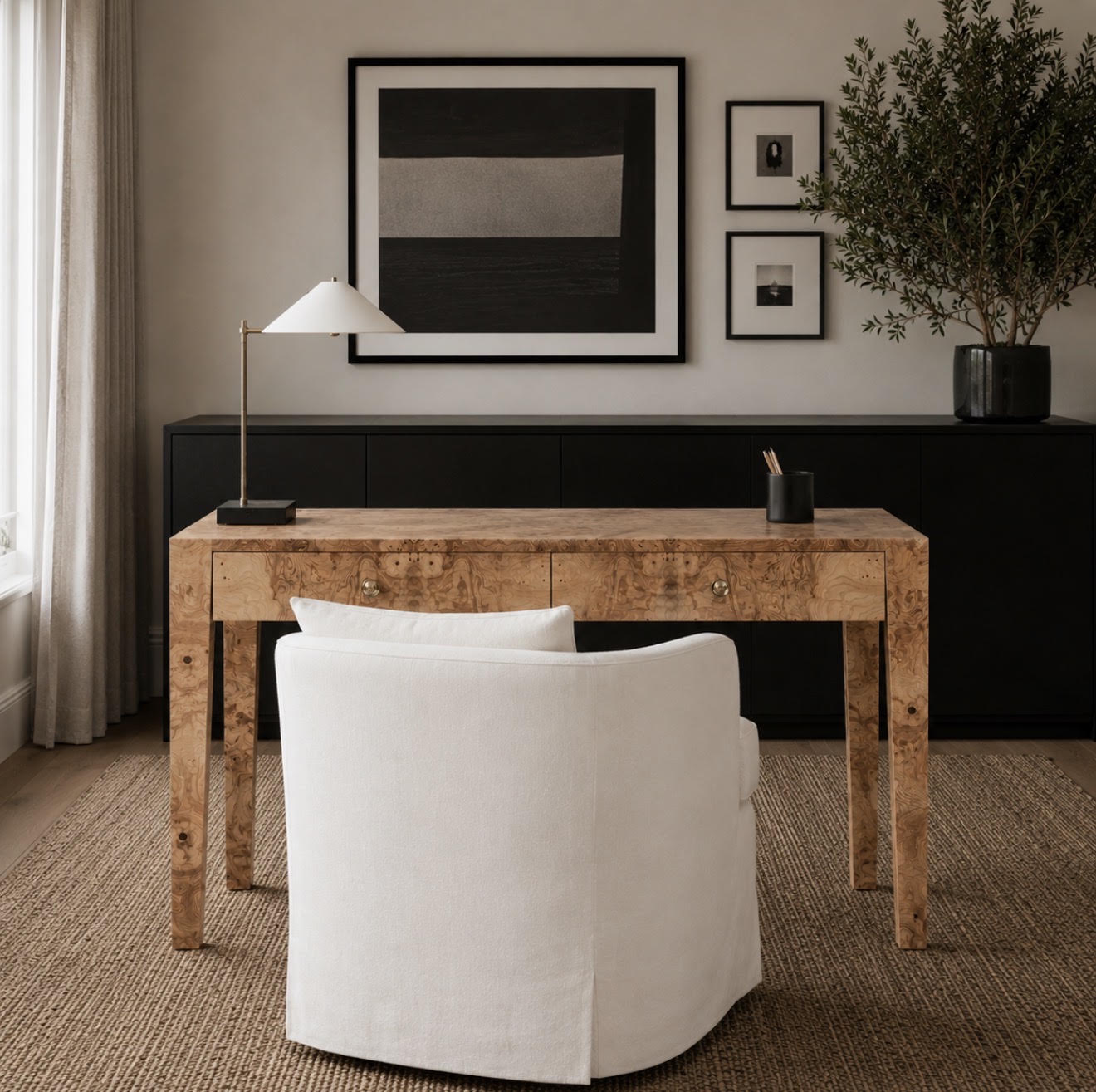 A rustic wooden writing desk with a white upholstered chair in a styled home office setting, featuring a black sideboard, framed wall art, and a table lamp.