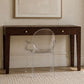 A dark brown wooden console table with two drawers and brass knobs, paired with a transparent acrylic armchair, set against a beige textured wall with a framed abstract print.