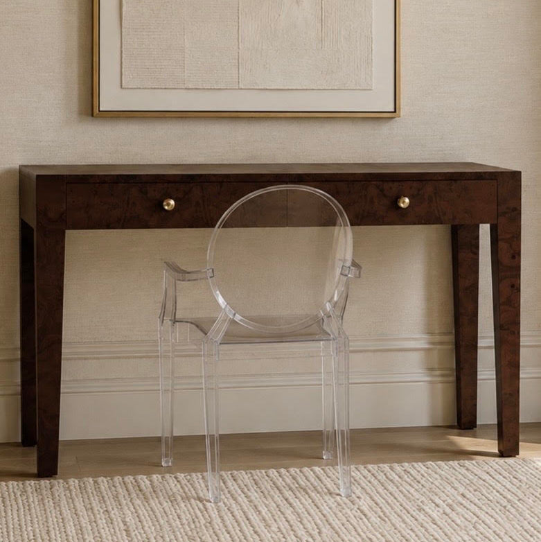 A dark brown wooden console table with two drawers and brass knobs, paired with a transparent acrylic armchair, set against a beige textured wall with a framed abstract print.