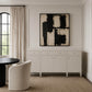 A large square abstract wall art print with black and off-white brushstrokes hangs above a white sideboard in a bright, modern room with beige curtains, a textured rug, and a cream upholstered chair.