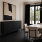 A modern dining room corner featuring a dark charcoal sideboard cabinet with brass knobs, a large abstract black and beige wall art piece, black-framed windows with dark curtains, and upholstered dining chairs.