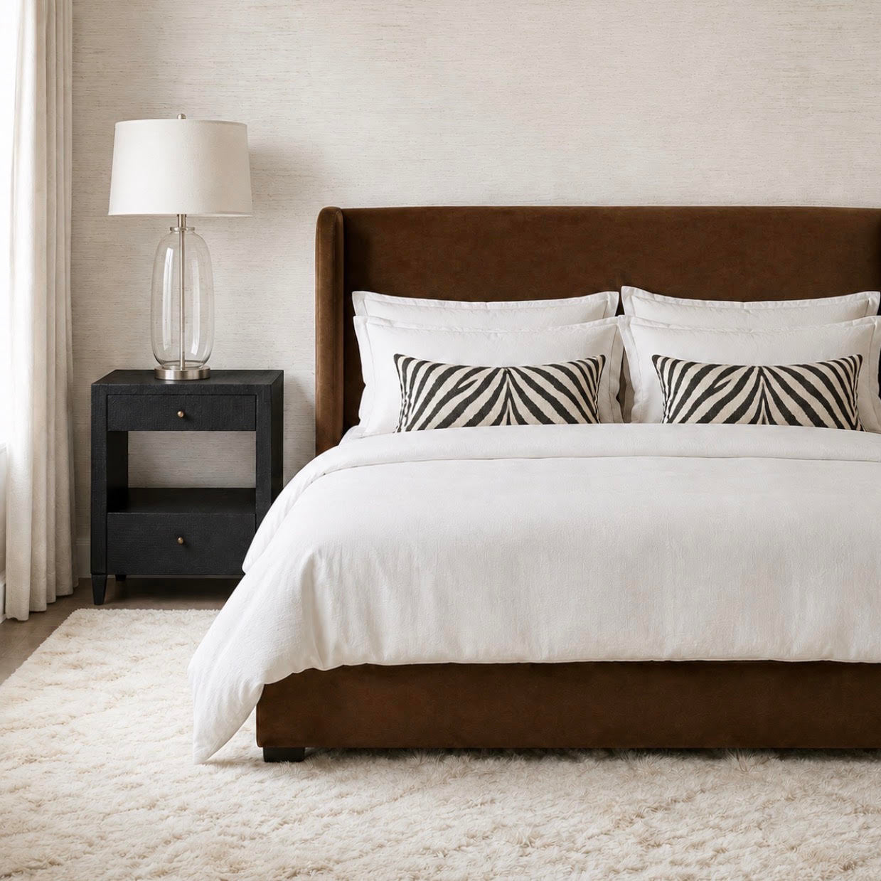 A styled bedroom scene featuring a bed with a brown upholstered headboard and matching footboard, dressed in white bedding with two black-and-white patterned accent pillows. A dark nightstand with a glass lamp sits beside the bed against a textured beige wall and white curtains.