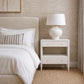 A modern bedroom scene featuring a white two-drawer nightstand with a white ceramic table lamp on top, positioned next to a bed with white linens and a striped accent pillow against a textured beige wall.