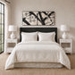 A neatly made bed with white bedding and two matching pillows, set in a bright bedroom with white nightstands, white lamps, a textured beige wall, and a black-framed abstract wall art piece above the headboard.