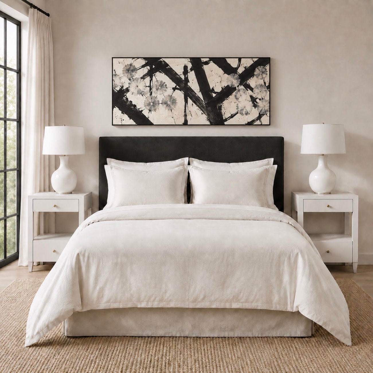 A neatly made bed with white bedding and two matching pillows, set in a bright bedroom with white nightstands, white lamps, a textured beige wall, and a black-framed abstract wall art piece above the headboard.