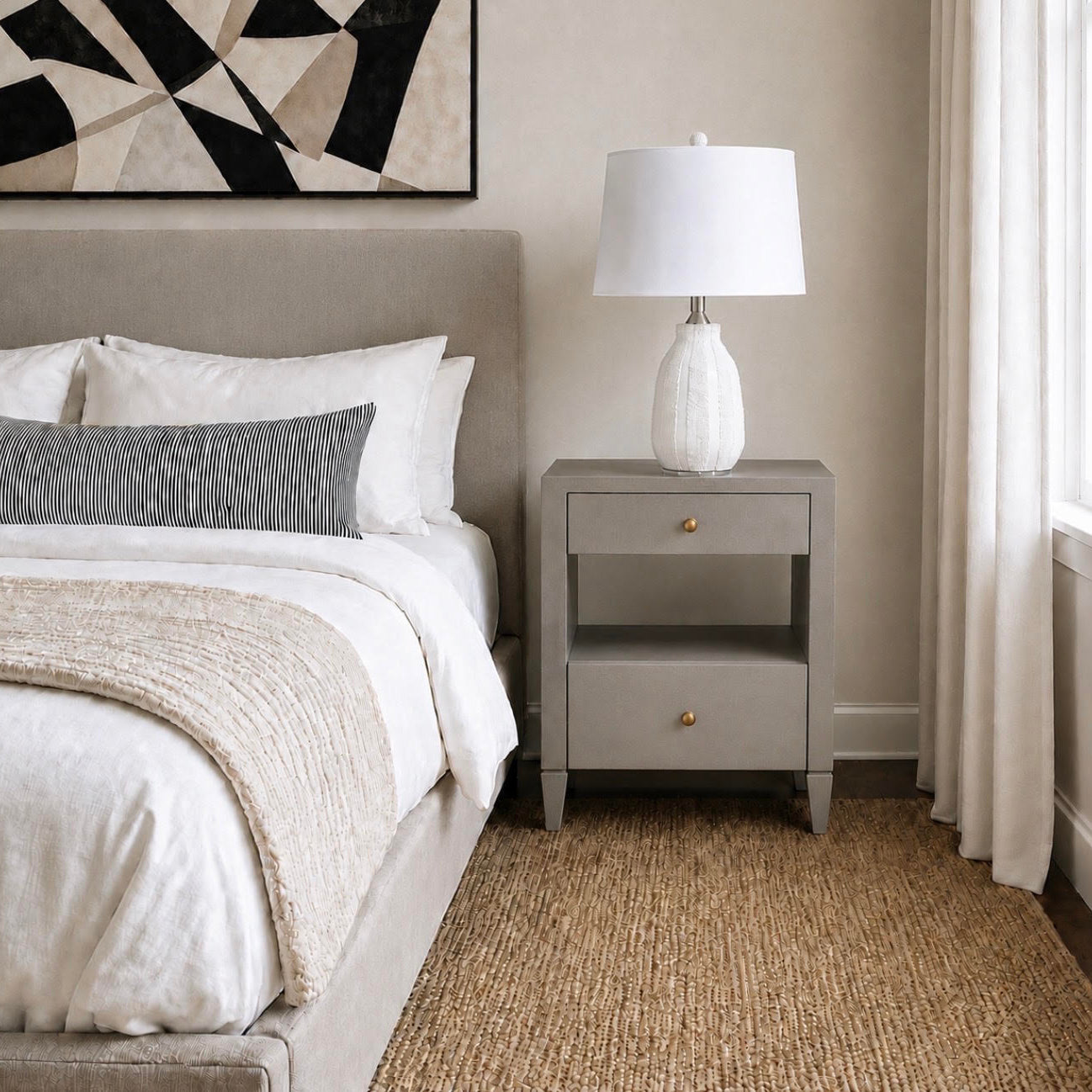 A styled bedroom scene featuring a light gray upholstered nightstand beside a bed with white linens and a textured throw blanket. A white ceramic table lamp sits on the nightstand, and a window with sheer curtains is visible on the right.