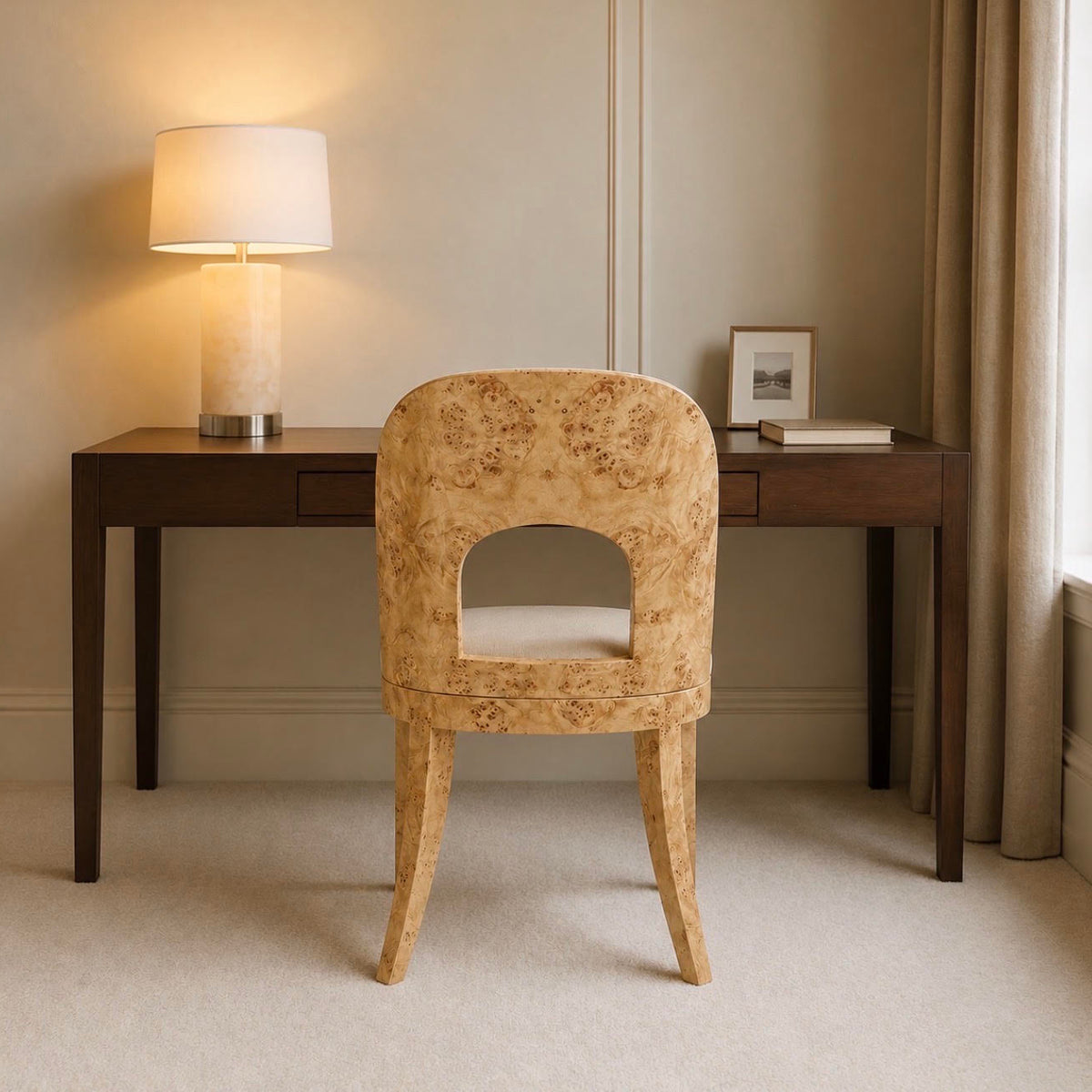 A beige upholstered accent chair with a cutout back, positioned in front of a dark wood writing desk in a softly lit room with a lit table lamp, curtains, and a window.