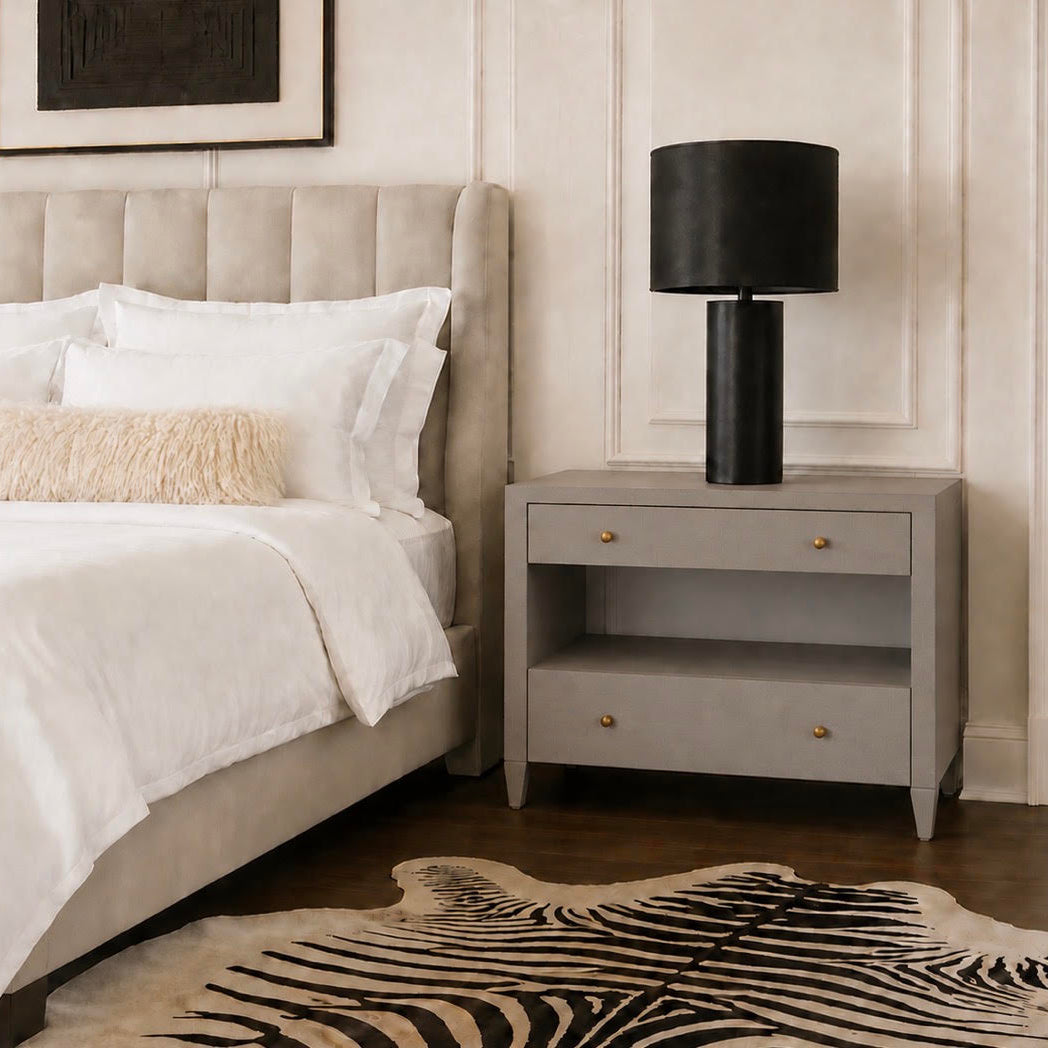 A modern bedroom scene featuring a light gray nightstand with a black table lamp, next to a bed with white linens and a textured cream pillow, set against a beige paneled wall.