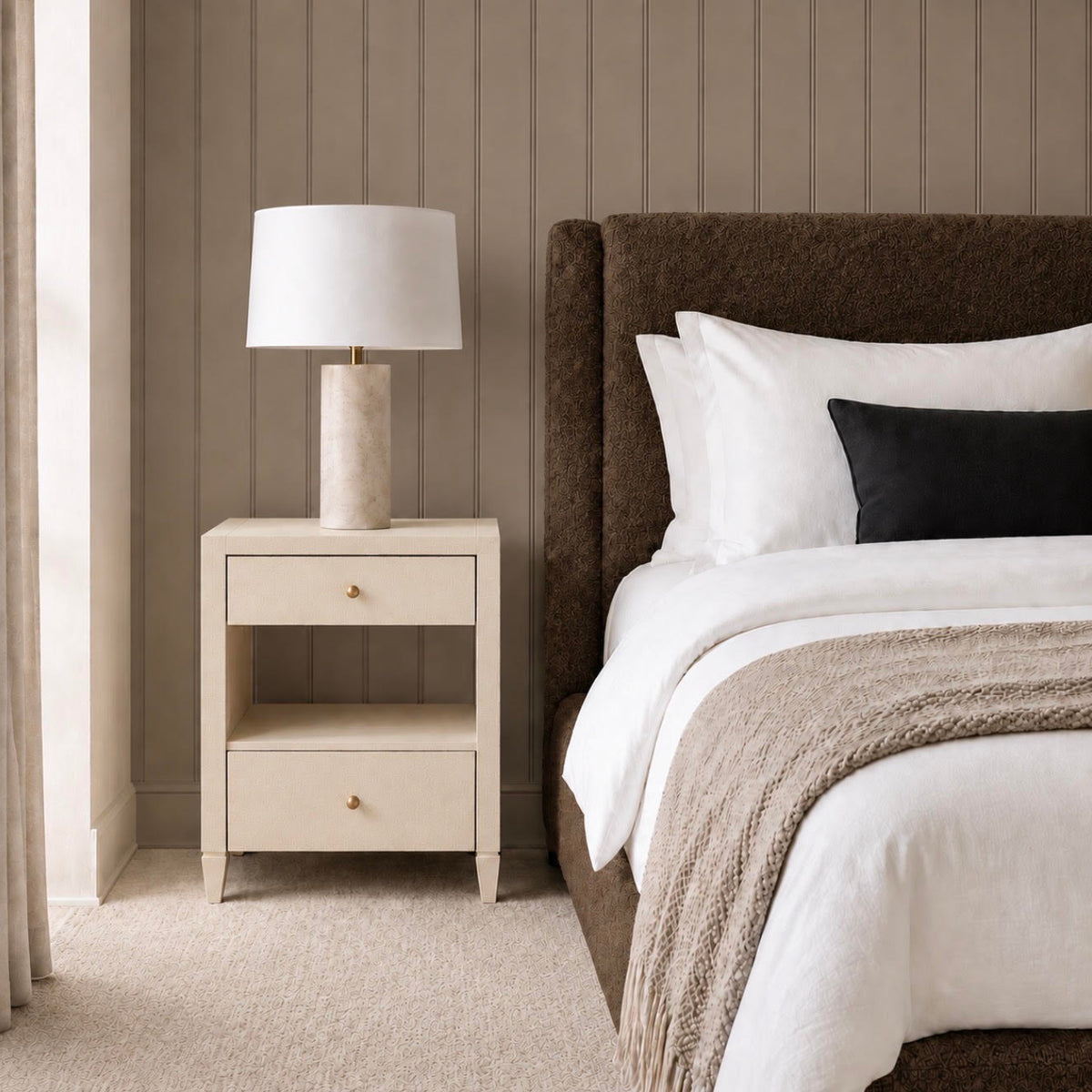 A styled bedroom scene featuring a cream-colored two-drawer nightstand with a textured stone lamp on top, positioned next to a bed with a dark brown upholstered headboard, white bedding, and a black accent pillow against a vertical wood-paneled wall.