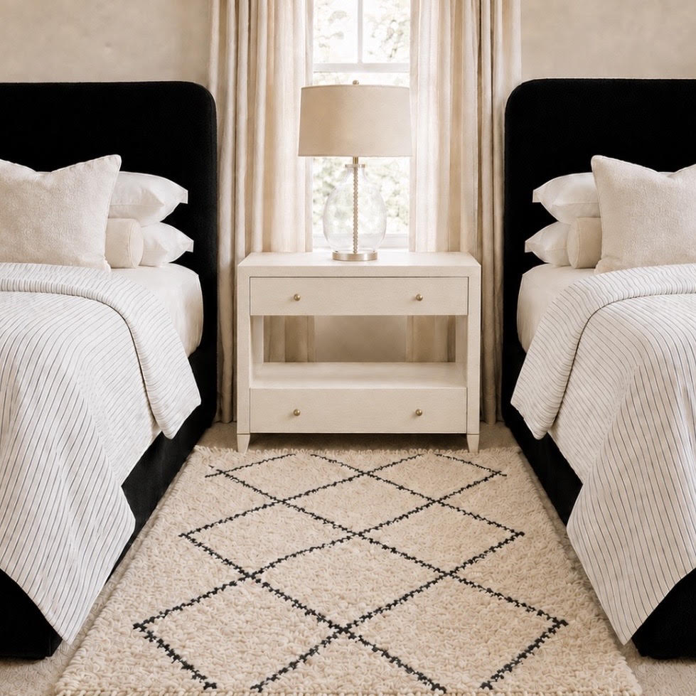 A styled bedroom scene with two twin beds, black upholstered heads, striped white bedding, a cream nightstand, and a beige rug with a black geometric diamond pattern. A window with sheer curtains and a table lamp sits between the beds.