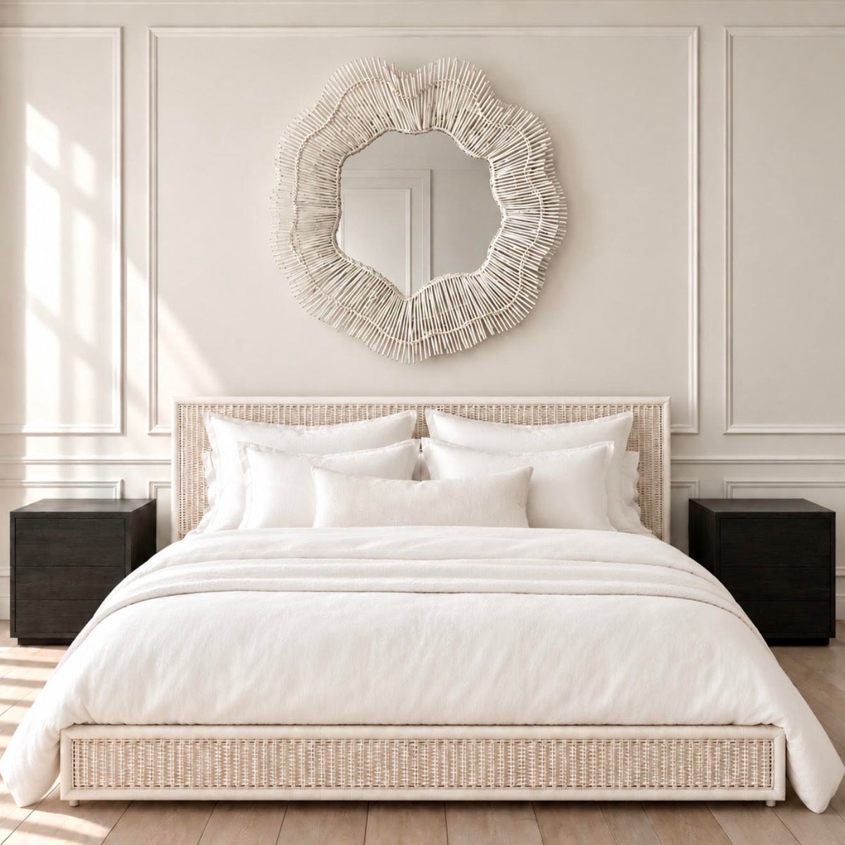 A styled bedroom scene featuring a bed with white bedding and pillows, a woven beige platform headboard and footboard, two dark nightstands, and a large round textured mirror mounted on a cream paneled wall.