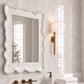 A large rectangular wall mirror with a textured, scalloped white frame hangs on a glossy white tiled bathroom wall above a marble sink. A gold-framed sconce, brass faucet hardware, white towels, and a soap dispenser are visible in the scene.