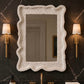 A large rectangular wall mirror with a heavily textured, pebbled off-white frame, mounted on a dark marble bathroom wall above a vanity with gold fixtures and flanked by two gold sconces.