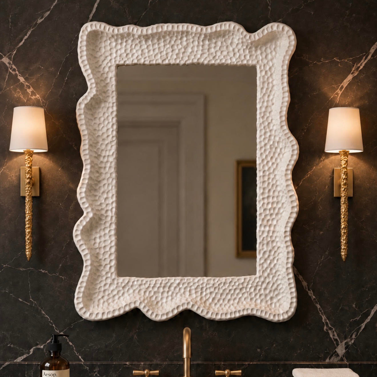 A large rectangular wall mirror with a heavily textured, pebbled off-white frame, mounted on a dark marble bathroom wall above a vanity with gold fixtures and flanked by two gold sconces.