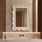 A rectangular bathroom mirror with a textured, pebbled white frame and scalloped edges, mounted on a marble-look wall above a matching marble sink vanity with warm wood accents.