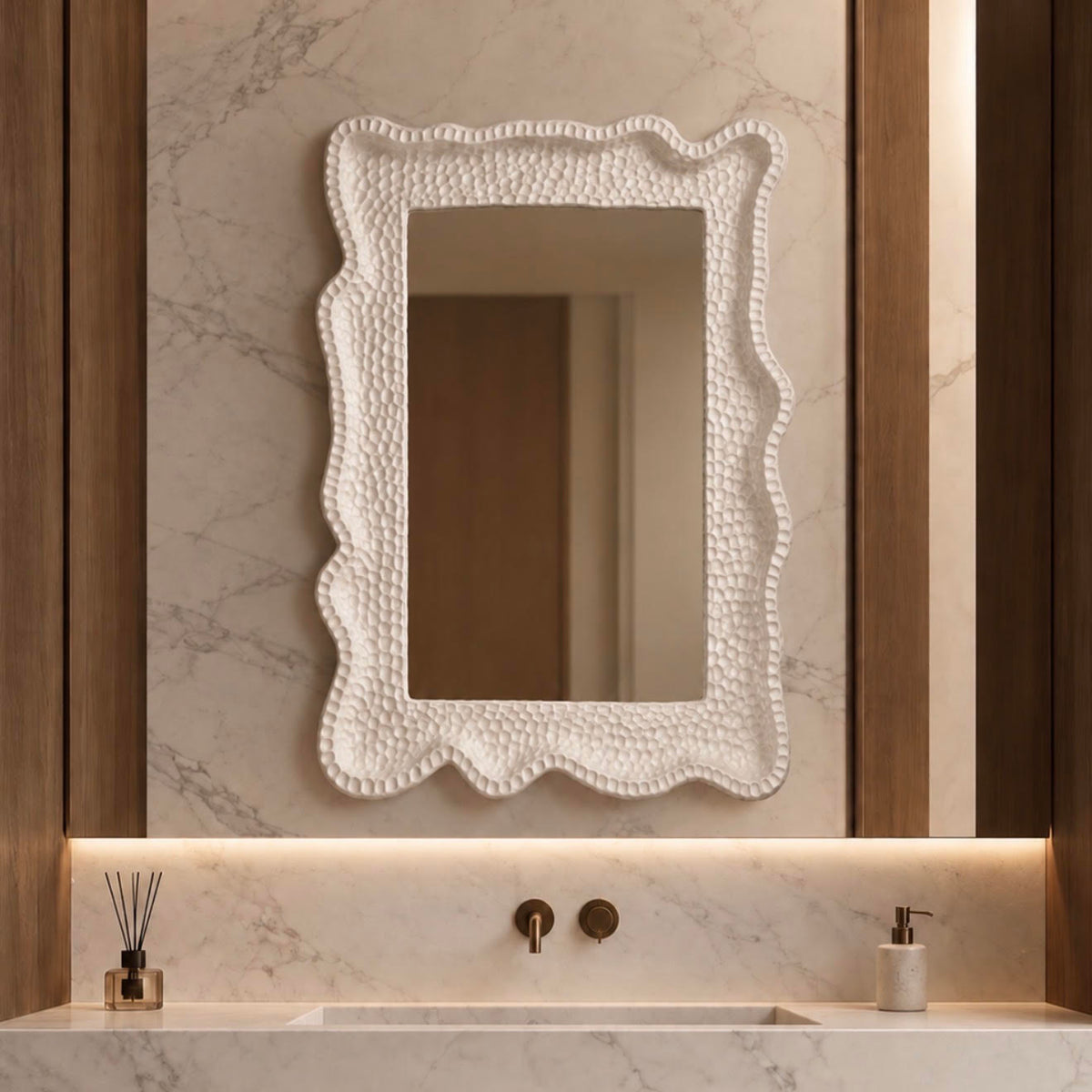 A rectangular bathroom mirror with a textured, pebbled white frame and scalloped edges, mounted on a marble-look wall above a matching marble sink vanity with warm wood accents.