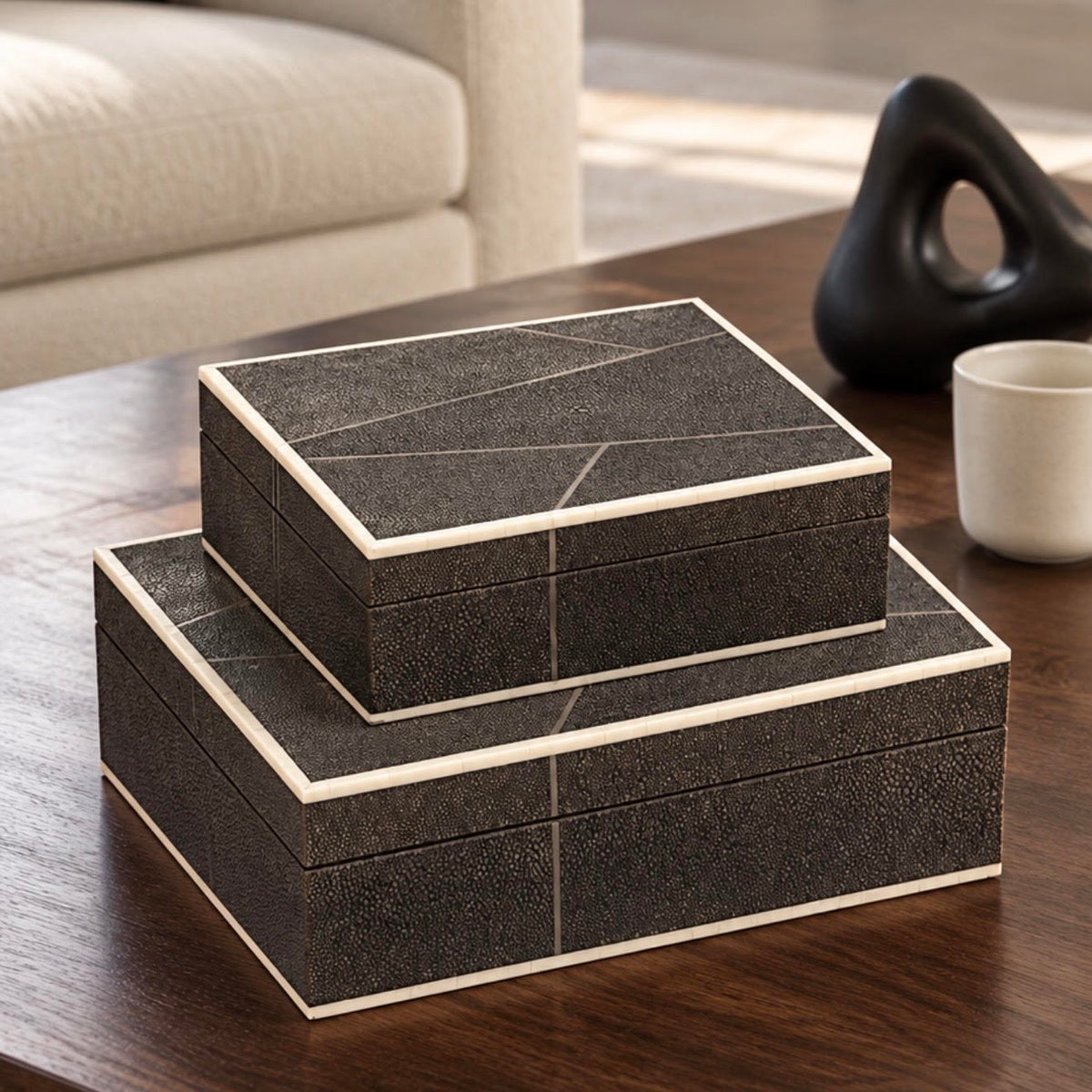 Two stacked rectangular storage boxes with a dark textured exterior and cream-colored edges, placed on a wooden table in a modern living room setting.