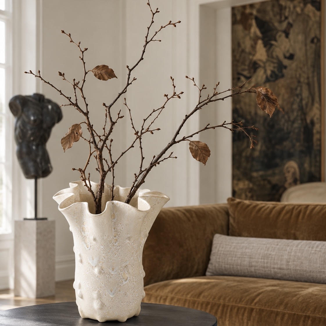 Sculptural ivory ceramic vase with an organic ruffled rim and textured finish, styled with dried autumn branches on a dark table in an elegant living room featuring a warm brown sofa, neutral accents, and vintage-inspired artwork.