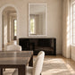 A large rectangular wall mirror with a wavy white frame hangs above a glossy black sideboard in a bright dining room. The room has beige textured walls, light wood floors, cream curtains, and a dark wood dining table with upholstered chairs.
