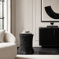 A black tree-stump side table styled in a modern living room setting next to a cream-colored armchair and a black ribbed credenza.