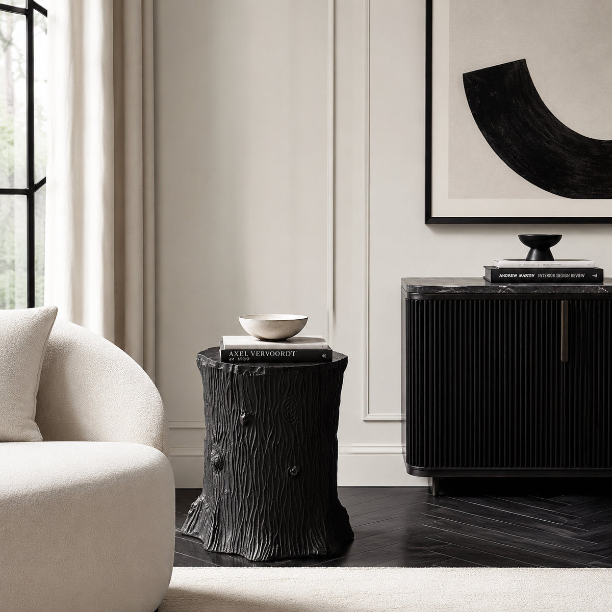 A black tree-stump side table styled in a modern living room setting next to a cream-colored armchair and a black ribbed credenza.