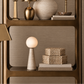 A styled interior scene showing a tall open shelving unit with decorative lamps, a vase, books, and woven boxes against a textured taupe wall. The shelving frame is a warm metallic gold/bronze tone, and the decor is arranged in a coordinated luxury setting.
