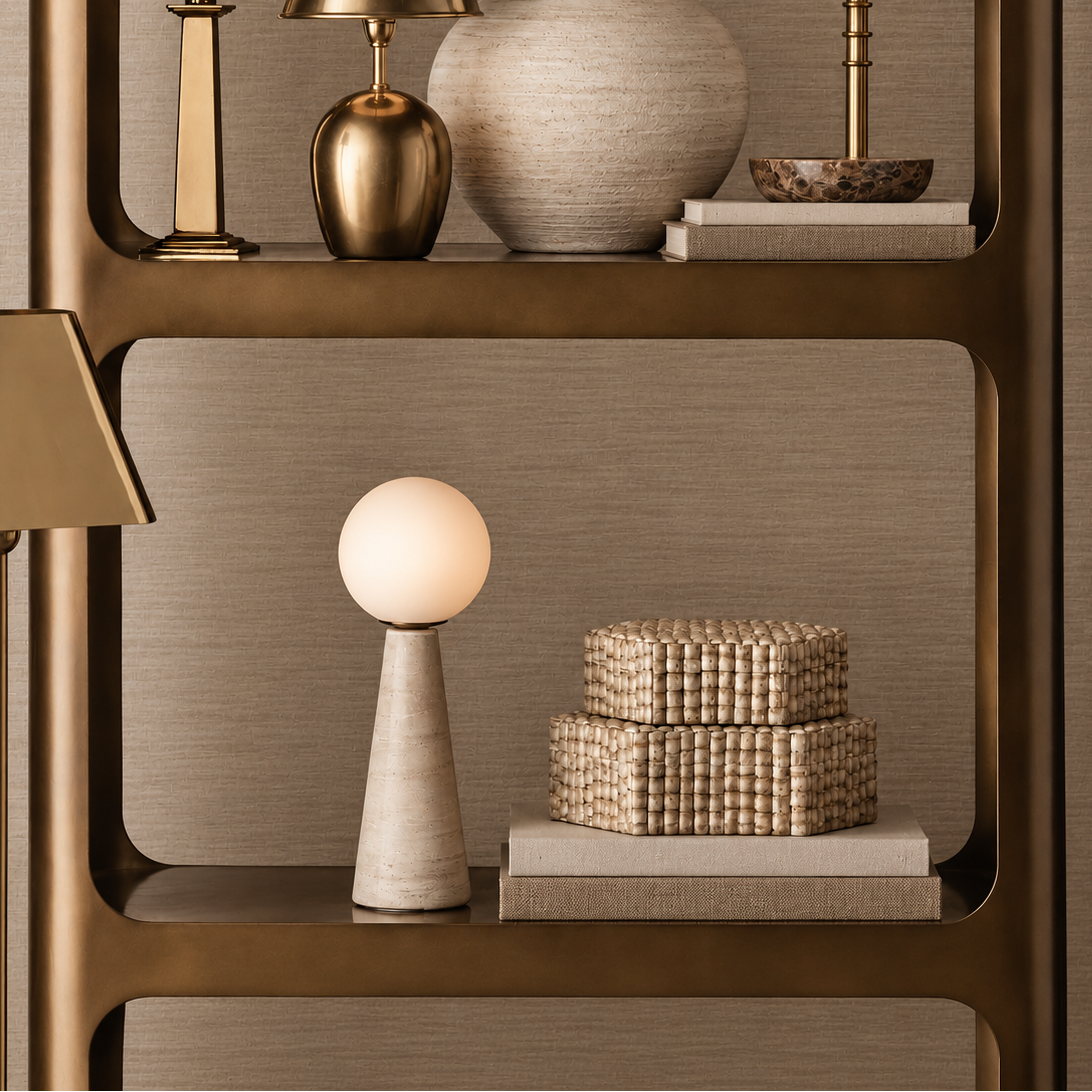 A styled interior scene showing a tall open shelving unit with decorative lamps, a vase, books, and woven boxes against a textured taupe wall. The shelving frame is a warm metallic gold/bronze tone, and the decor is arranged in a coordinated luxury setting.