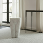 A modern white marble pedestal stool with subtle gray veining stands on a textured light gray rug in a bright, contemporary room with sheer curtains and a dark-framed window.
