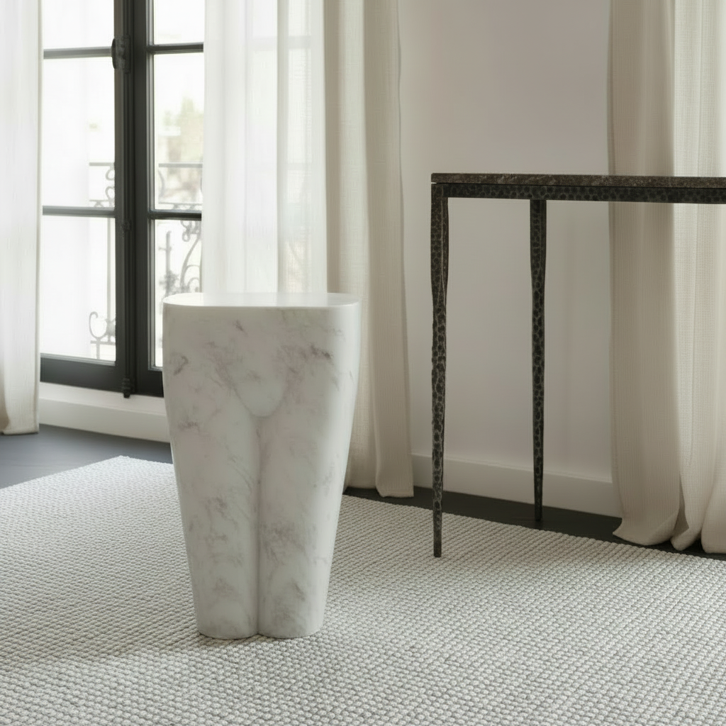 A modern white marble pedestal stool with subtle gray veining stands on a textured light gray rug in a bright, contemporary room with sheer curtains and a dark-framed window.