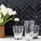 White tulips in a wooden vase with etched glass tumblers on a black tiled wall background