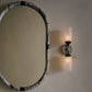 Round mirror with decorative frame on a wall next to a sconce.