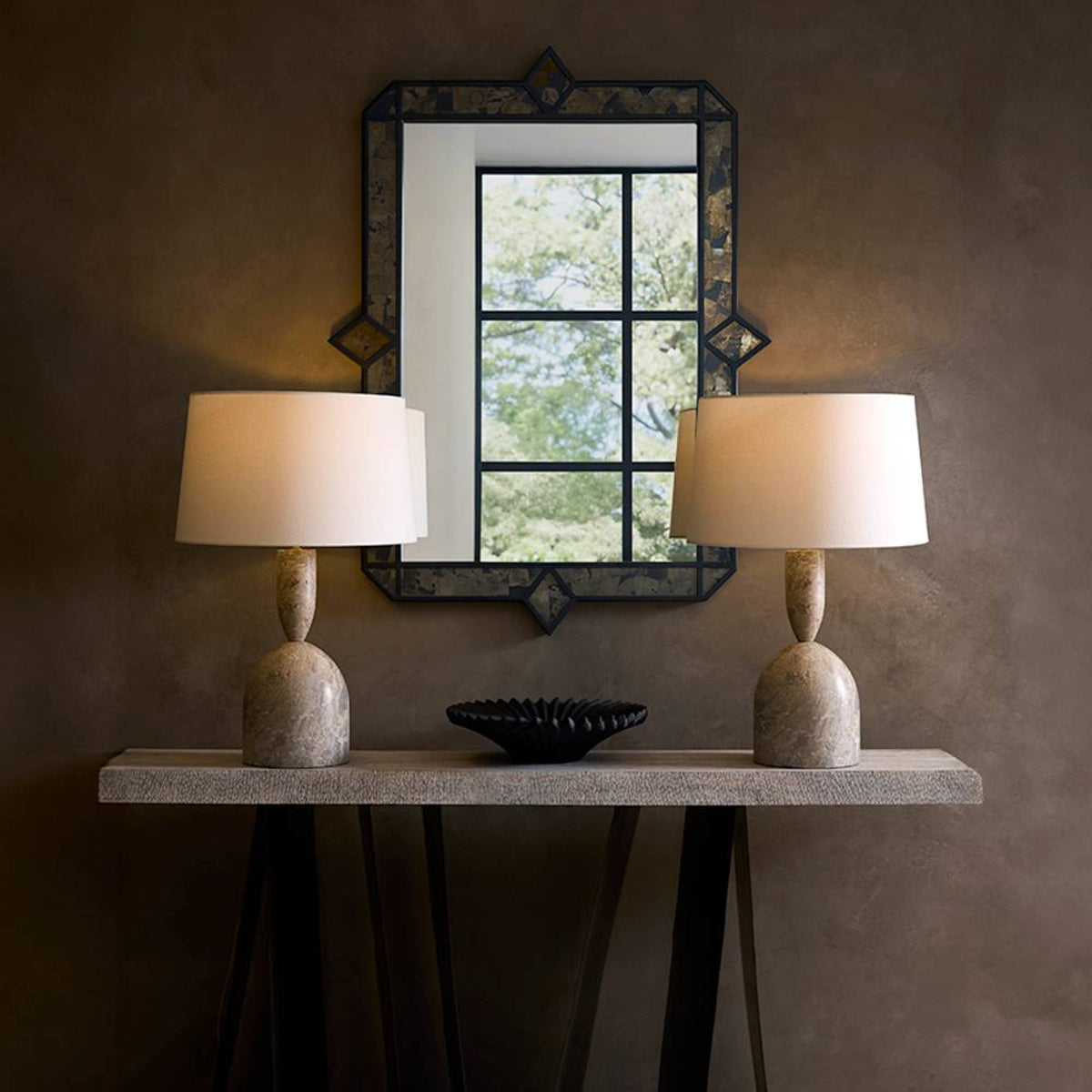 Two table lamps on a stand with a decorative mirror above them against a textured wall.